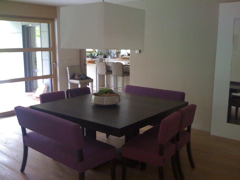 Salle à manger en retrait, table carrée en bois noir, chaises violettes. 