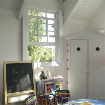 Chambre enfant avec poutres apparentes blanches. Table ronde en plastique bleu. Tableau ardoise et armoire pour enfant. Murs en bois blanc 